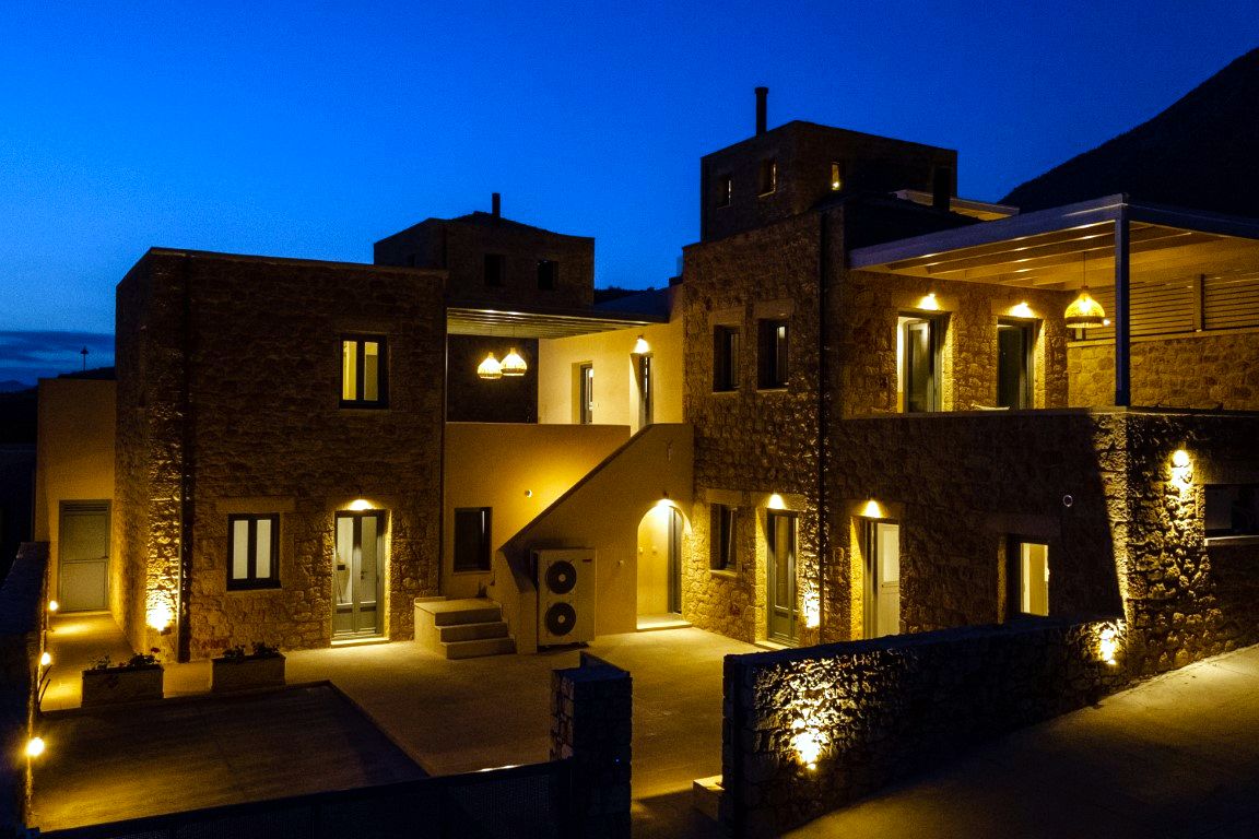 Two-story stone villas with multiple windows and balconies are lit by warm exterior lights against a deep blue twilight sky. A paved walkway leads between the structures.