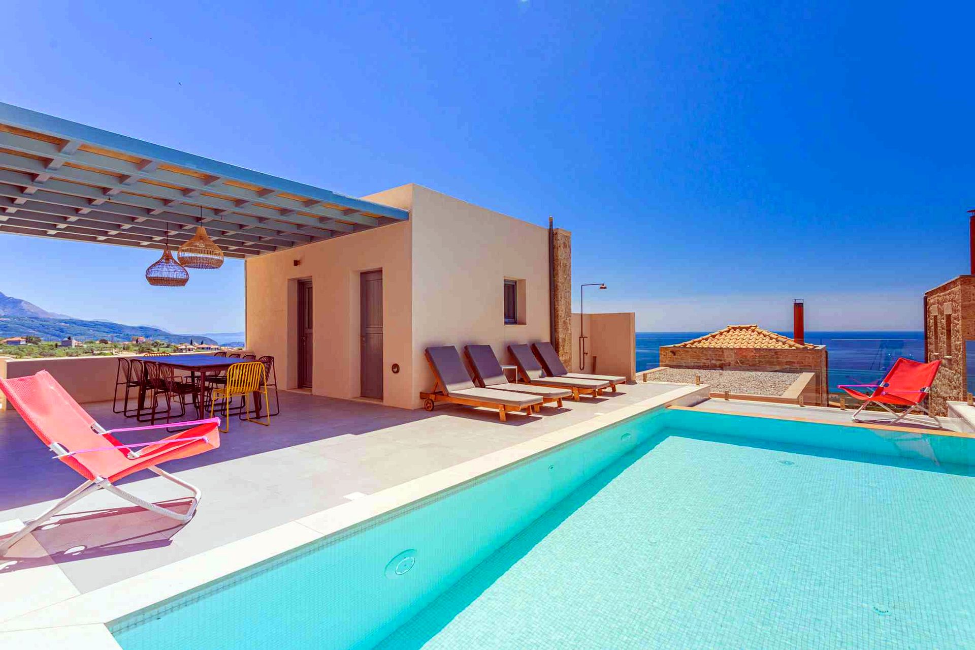 {
  "headline": "Poolside view from private villa patio",
  "subheadline": "Loungers and dining set sit beneath a shaded pergola overlooking the Aegean Sea.",
  "alt_text": "A rectangular swimming pool occupies the foreground, bordered by a light-colored patio. To the left, a pergola covers an outdo