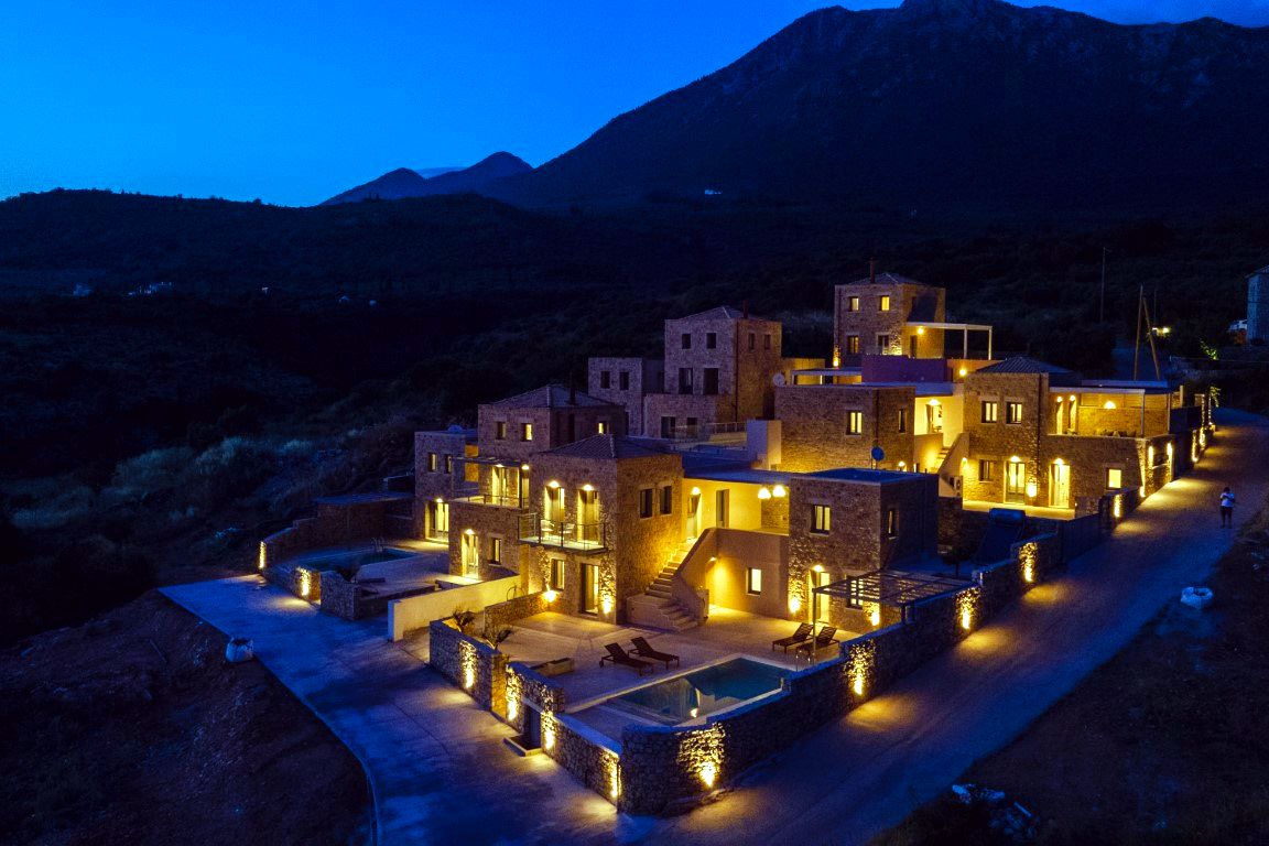 Group of stone villas with multiple levels and terracotta roofs are lit by exterior lamps at dusk. A rectangular pool is visible in the foreground courtyard area.