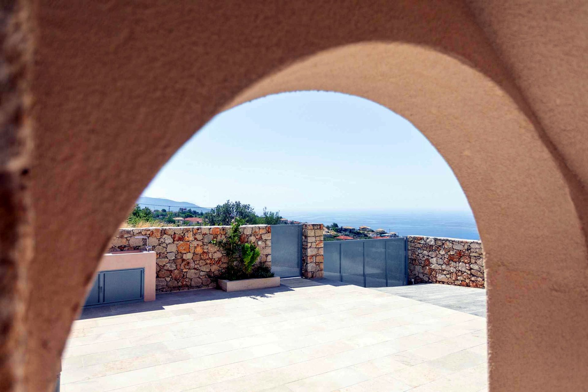 Stone patio area framed by a large terracotta archway overlooks a bright blue sea under a clear sky. Low stone walls and modern gray paneling line the edge of the space.