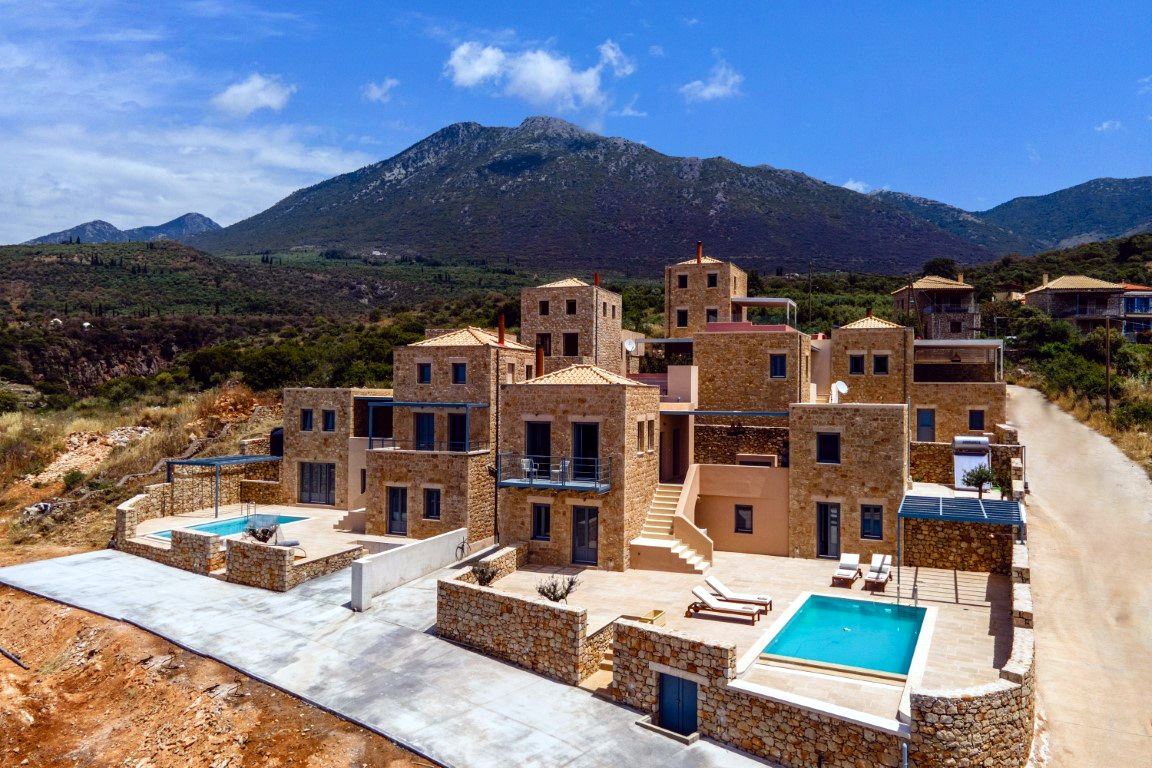 Group of multi-story stone villas with tiled roofs and balconies overlooks a valley with distant mountains under a bright blue sky. Several patios feature rectangular swimming pools.