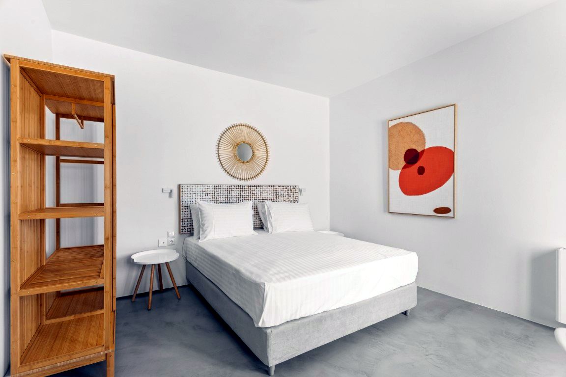 A bed with white linens and a woven circular headboard sits in a room with light gray floors. To the left, a tall wooden shelving unit stands against the wall. Artwork featuring red and orange circles hangs on the far wall.