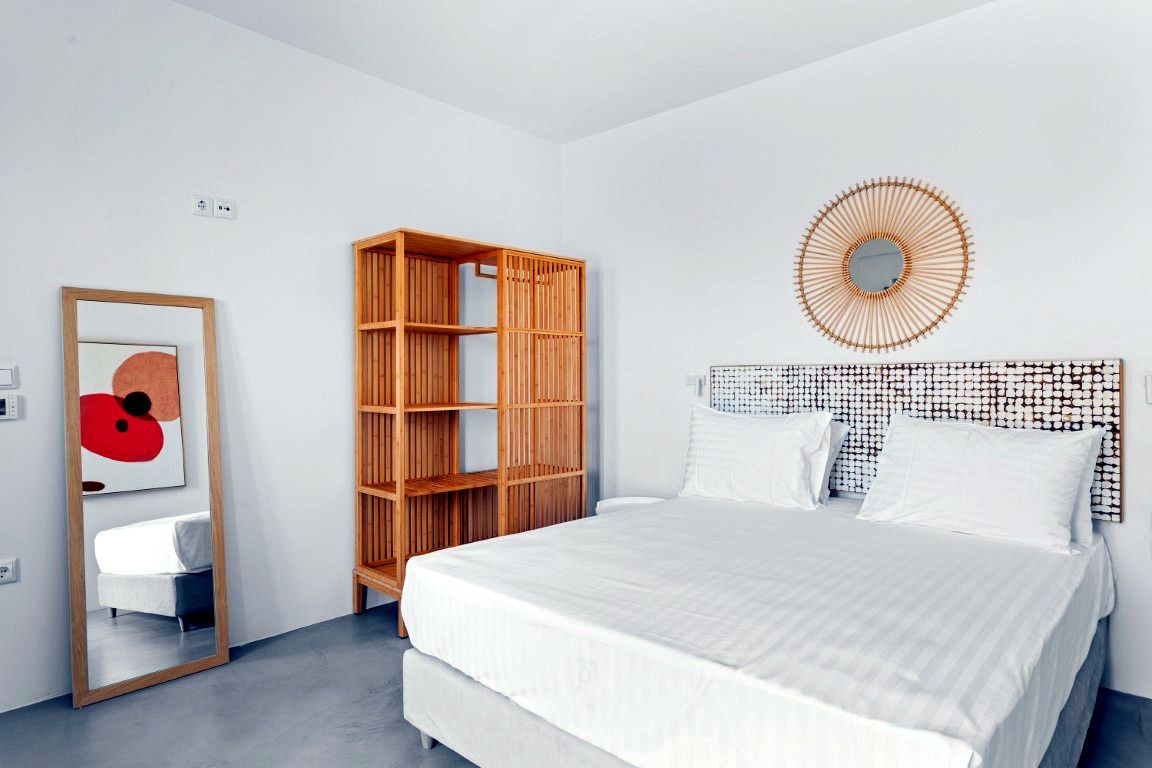 White bed with white linens occupies the foreground. To the left, a tall, open wooden shelving unit stands against the wall. Above the bed, a circular, patterned mirror hangs over a textured headboard.