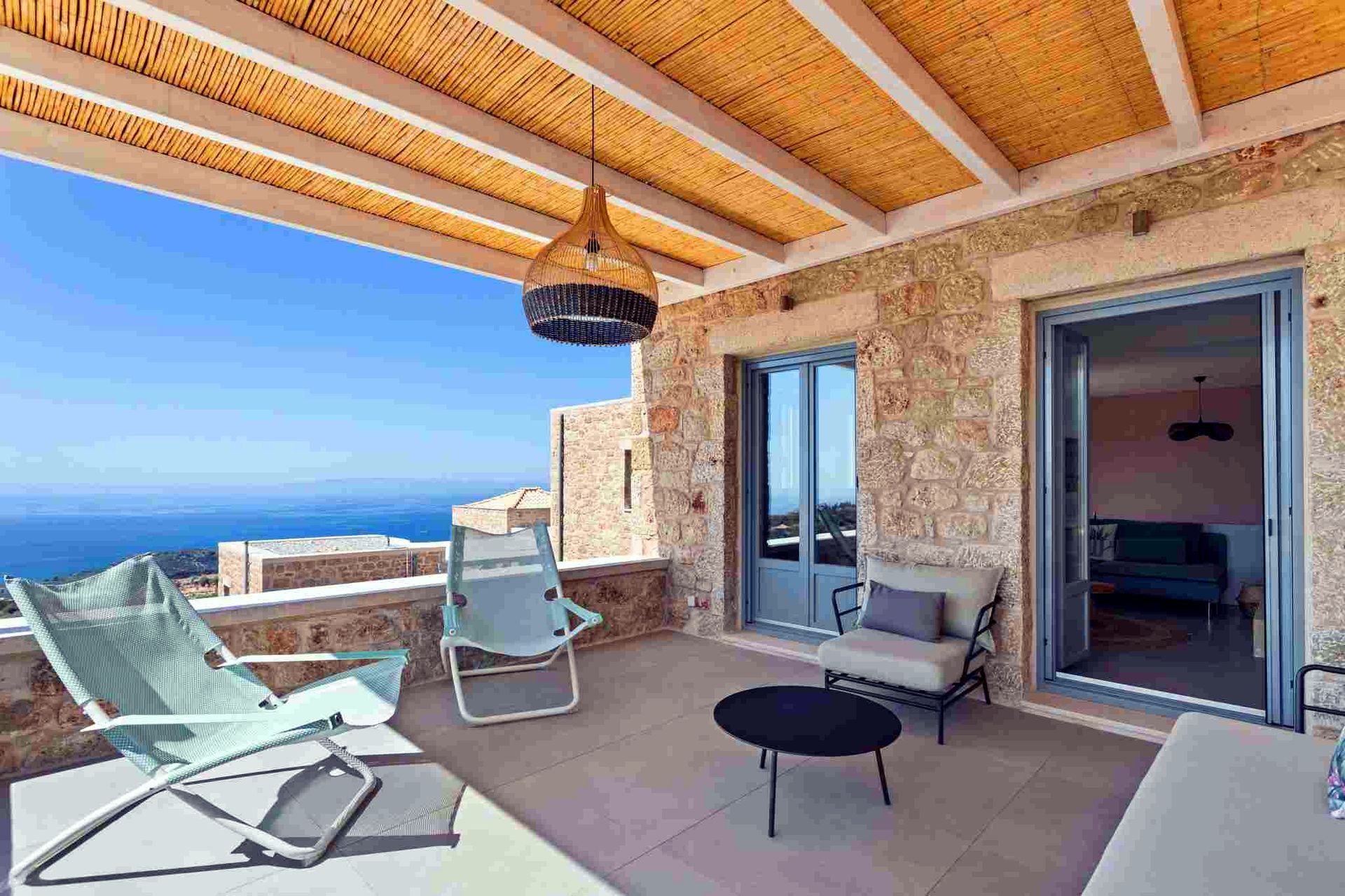 Two light green lounge chairs with adjustable backs sit on a tiled patio. The patio has a wooden slat ceiling and opens onto a view of the blue sea under a bright sky. A black circular table rests near the entrance.