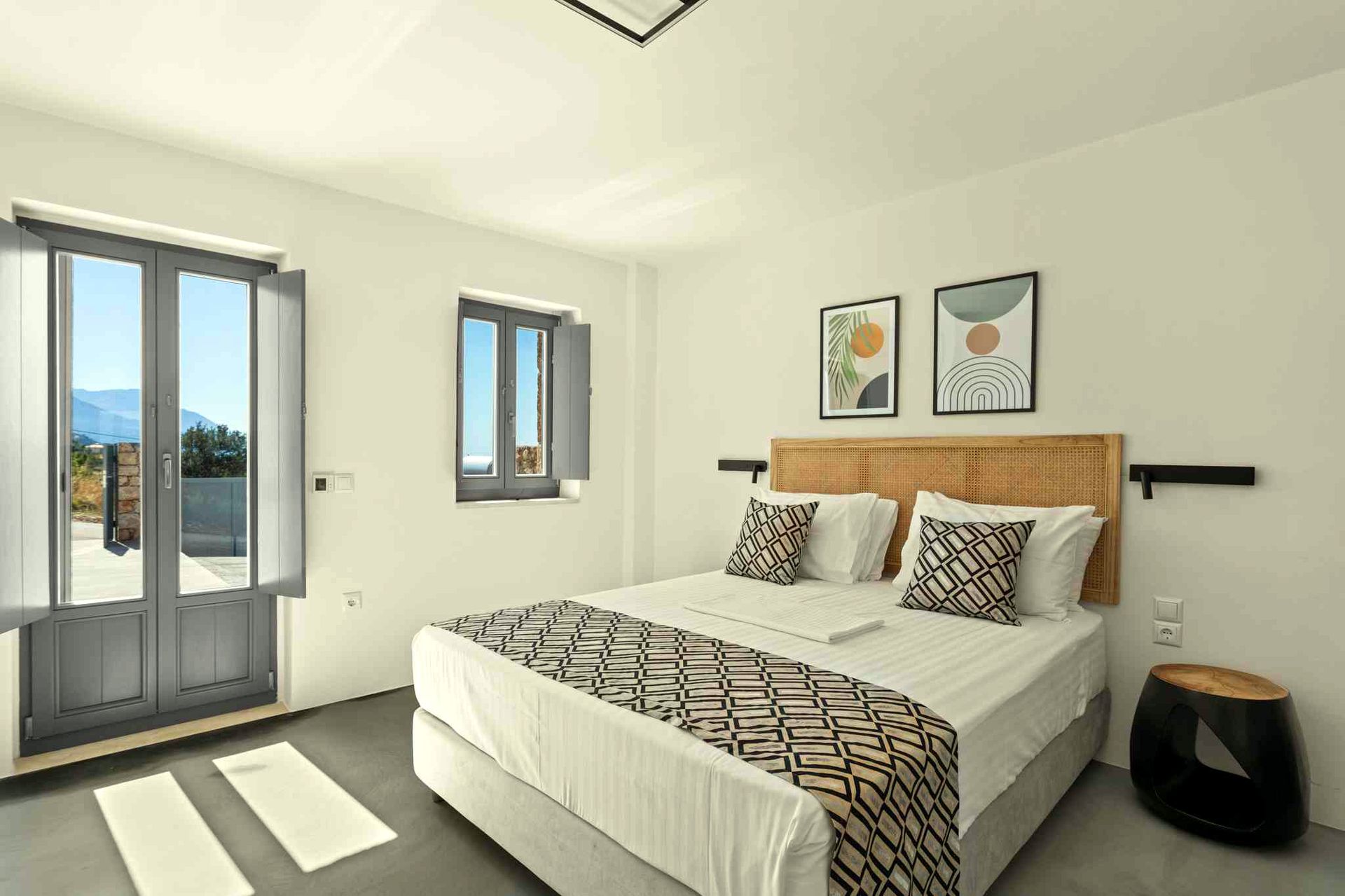 Two sets of sliding glass doors open onto a balcony overlooking a view of the sea and distant hills. Inside, a bed with a patterned throw rests against a wooden headboard. Two framed prints hang on the wall above the bed.