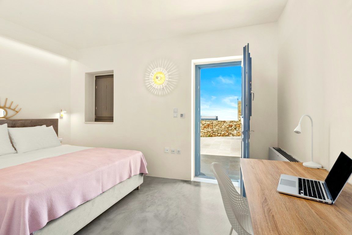 A bedroom interior with a large bed covered in pink linens, a wooden desk surface, and a doorway opening onto a bright blue exterior view.