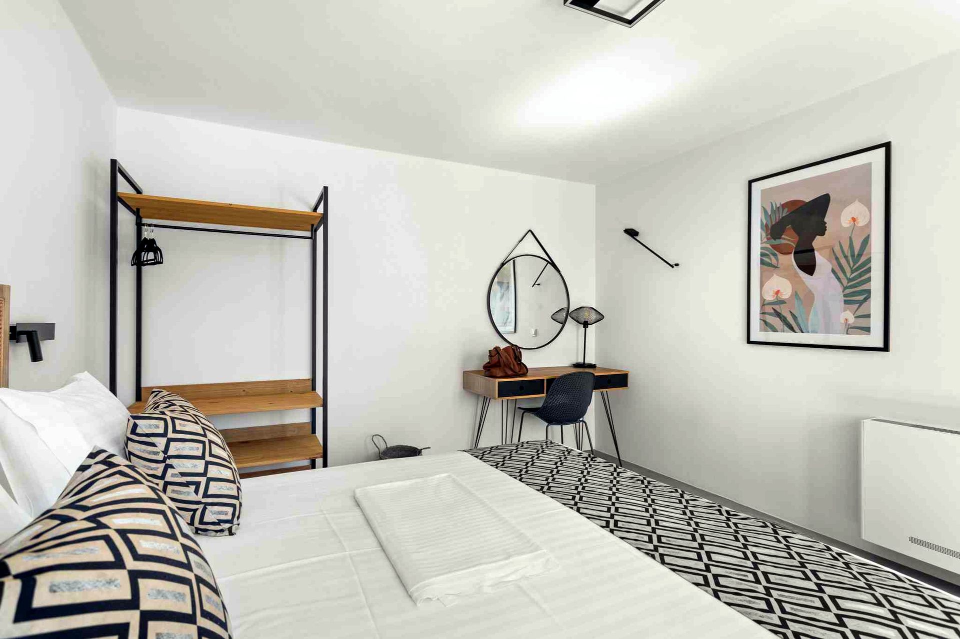 Black metal bed frame with wooden ladder shelf stands against a white wall. A round mirror hangs above a wooden desk with a black chair. A framed artwork hangs on the adjacent wall.