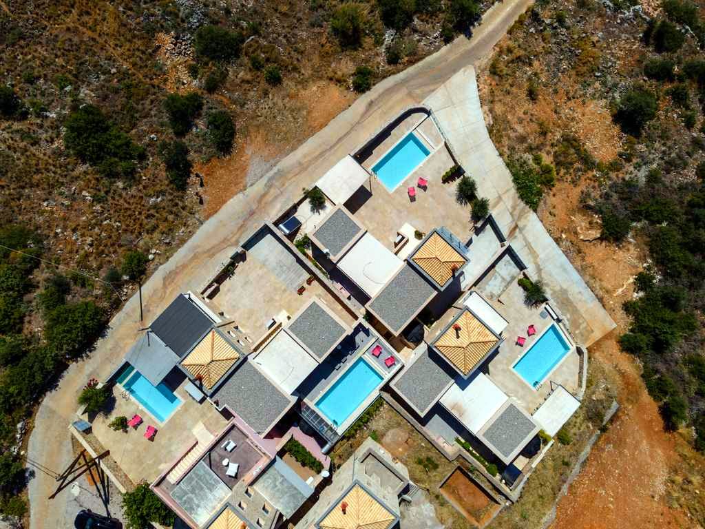 Aerial view of several attached villas with rectangular swimming pools and light-colored stone patios. The buildings are clustered together, surrounded by dry, scrubby vegetation and dirt pathways.