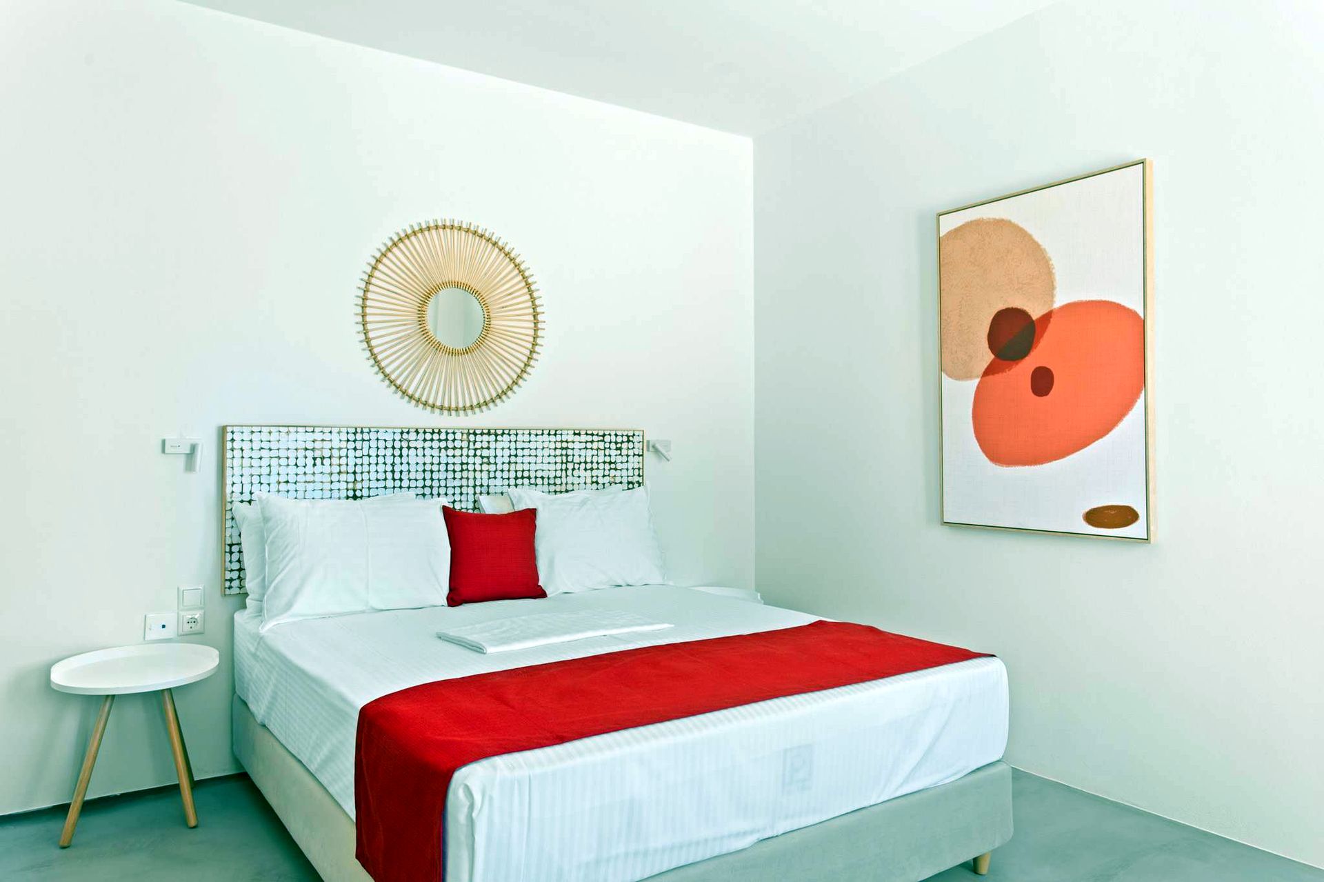 A bed with white linens and a red throw is centered in a room. Above the headboard, a circular woven mirror hangs. To the right, a piece of artwork features abstract orange and white shapes.