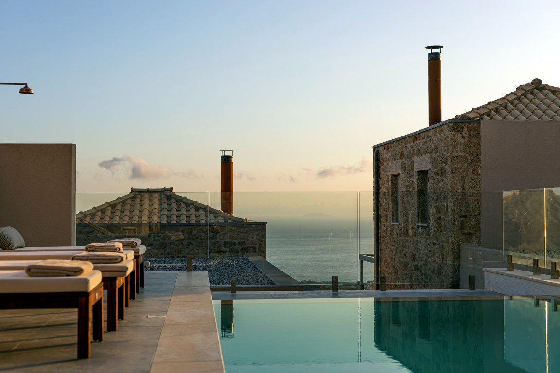 A rectangular swimming pool sits in the foreground, bordered by light-colored paving stones. To the right is a stone building with a tiled roof and vertical chimney stacks. The background shows a vast expanse of water meeting a pale sky.