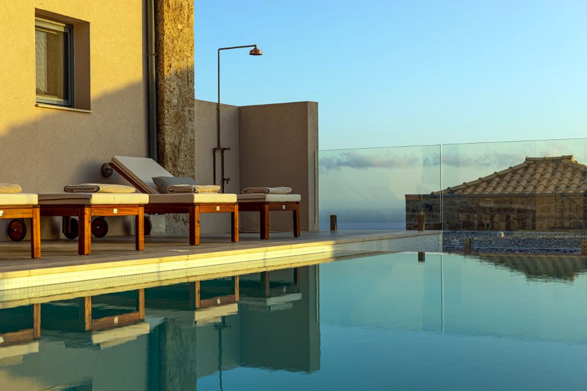 Two wooden lounge chairs with white cushions sit on a tiled patio next to a building wall. A glass railing separates the patio from a view of the sea and a low-profile structure.