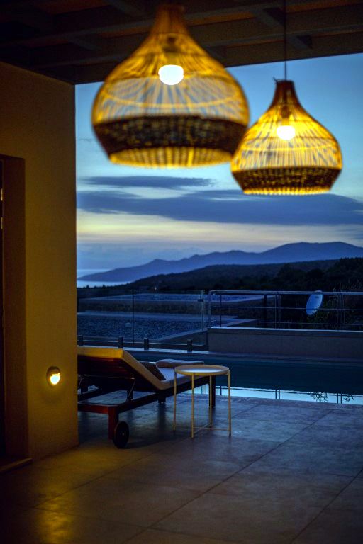 Two large, woven pendant lights hang from a covered patio structure. Below the lights, a low wooden lounge chair rests on a tiled floor, overlooking a dark expanse of water and distant hills under a twilight sky.