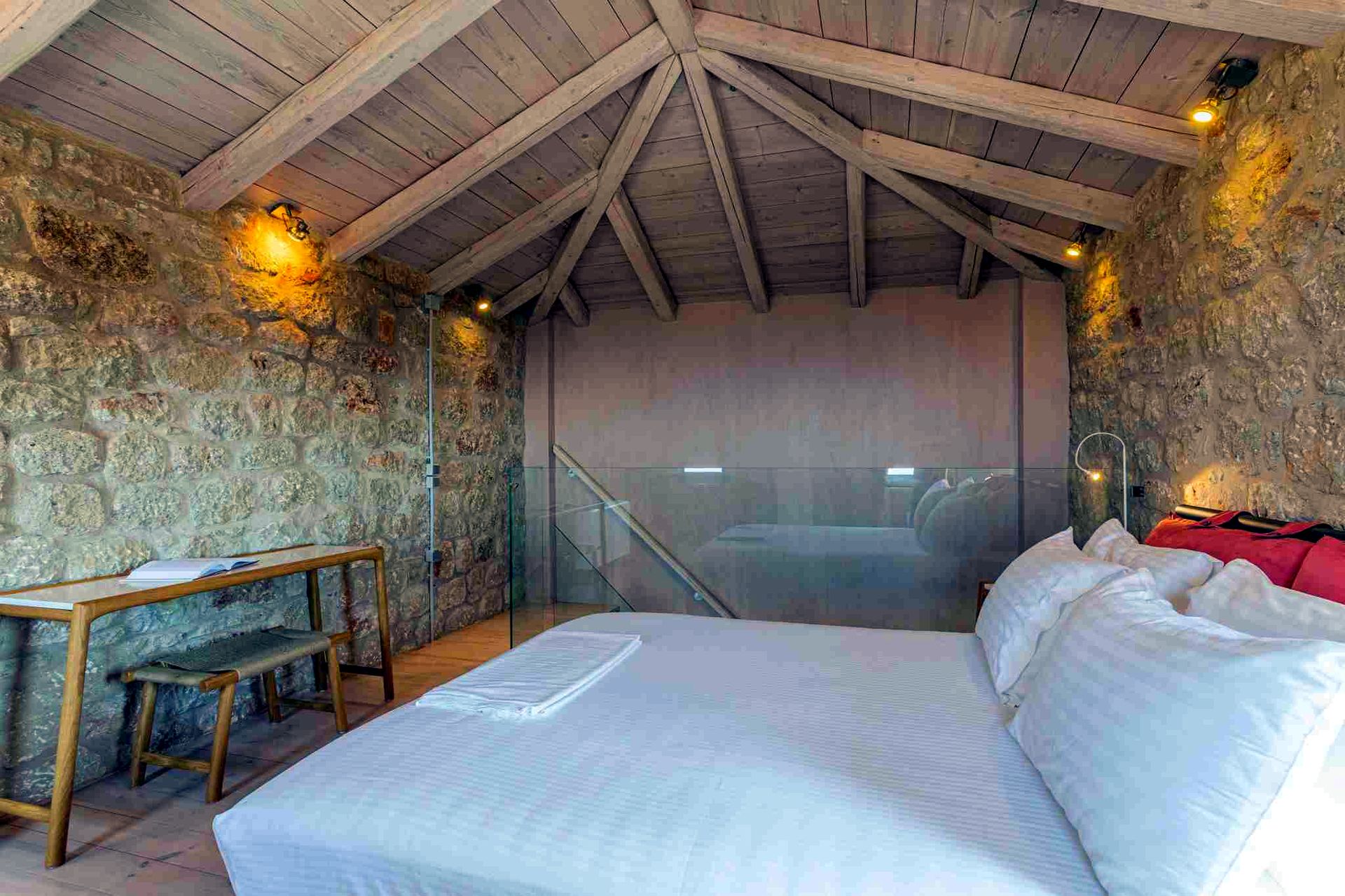 A bedroom with exposed wooden beams on the ceiling and rough stone walls. A low wooden bench rests against the left wall, and a bed with white linens occupies the center.