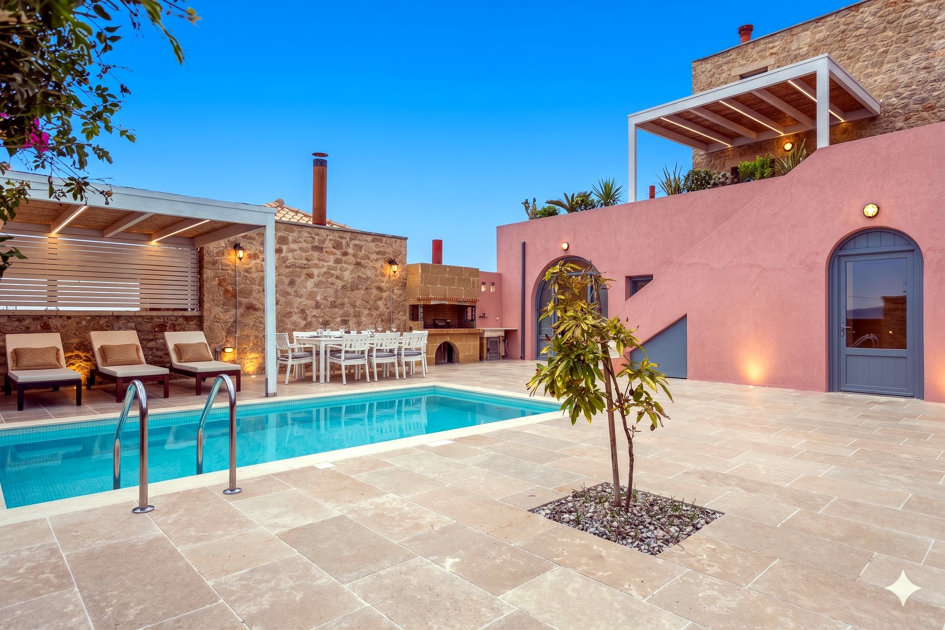Rectangular swimming pool occupies the foreground patio area. To the left, a covered outdoor dining space is visible. The background features a pink stucco building with a recessed doorway and a stone-clad section with a balcony.