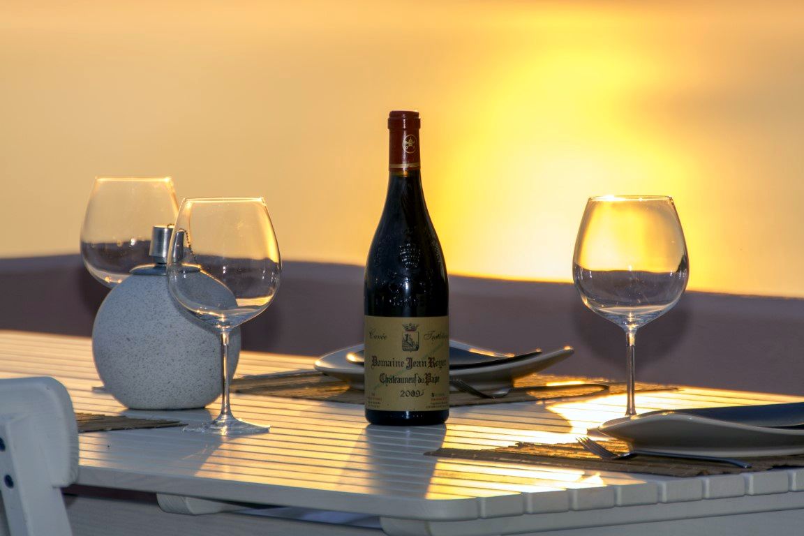 A dark green wine bottle stands on a white table surface. To the left, a speckled white vase sits next to two empty wine glasses. To the right, a single empty wine glass is visible.