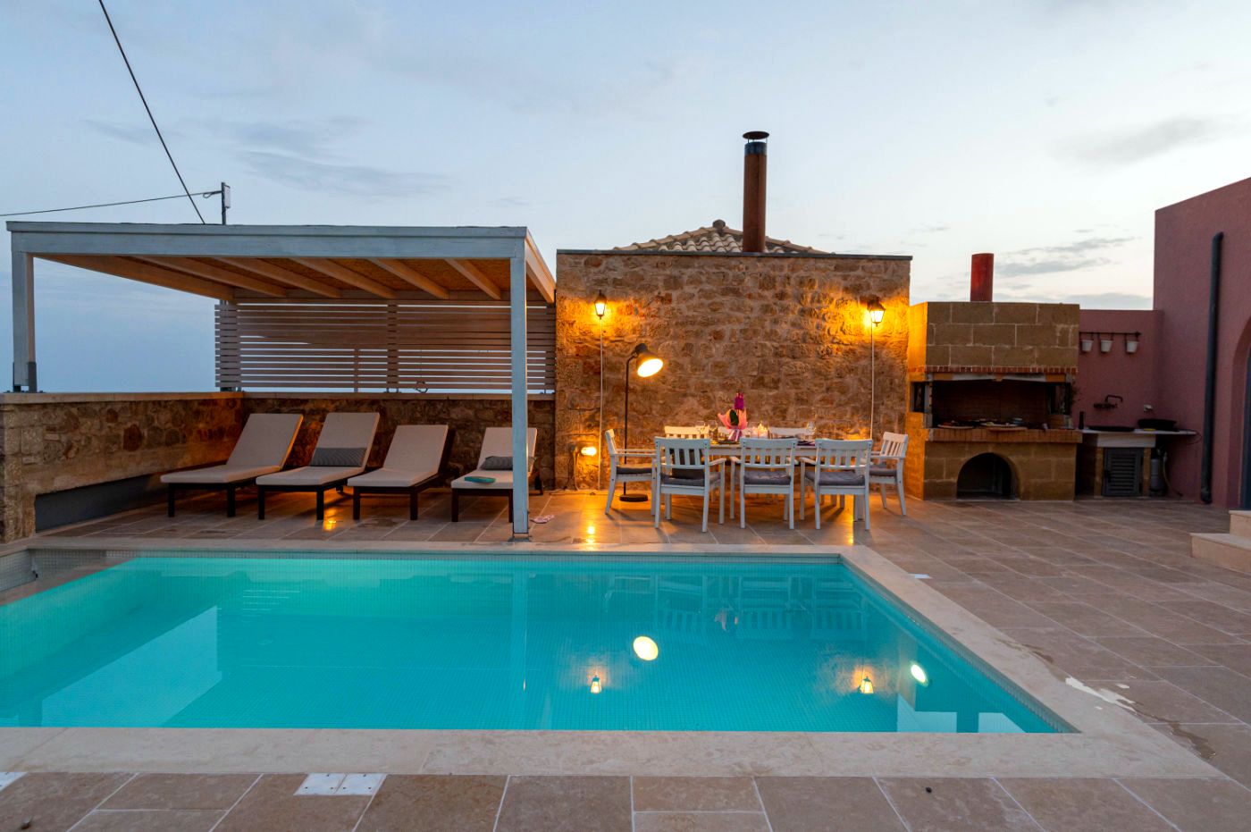Rectangular swimming pool occupies the foreground, bordered by light tile. Behind the pool is a covered patio area with wooden slats. To the right stands a stone building with a chimney and an outdoor grill area.