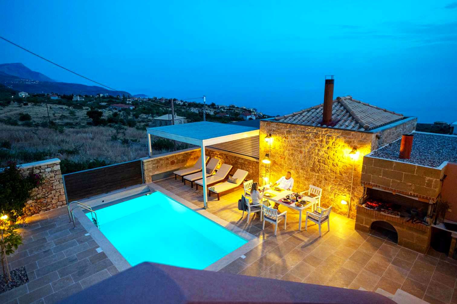 {
  "headline": "Pool overlooks Aegean Sea at Dusk",
  "subheadline": "Stone patio surrounds the rectangular pool, with a visible outdoor dining set near the building's corner.",
  "alt_text": "Rectangular swimming pool set into a stone patio area, viewed at dusk. The pool water is bright blue, and