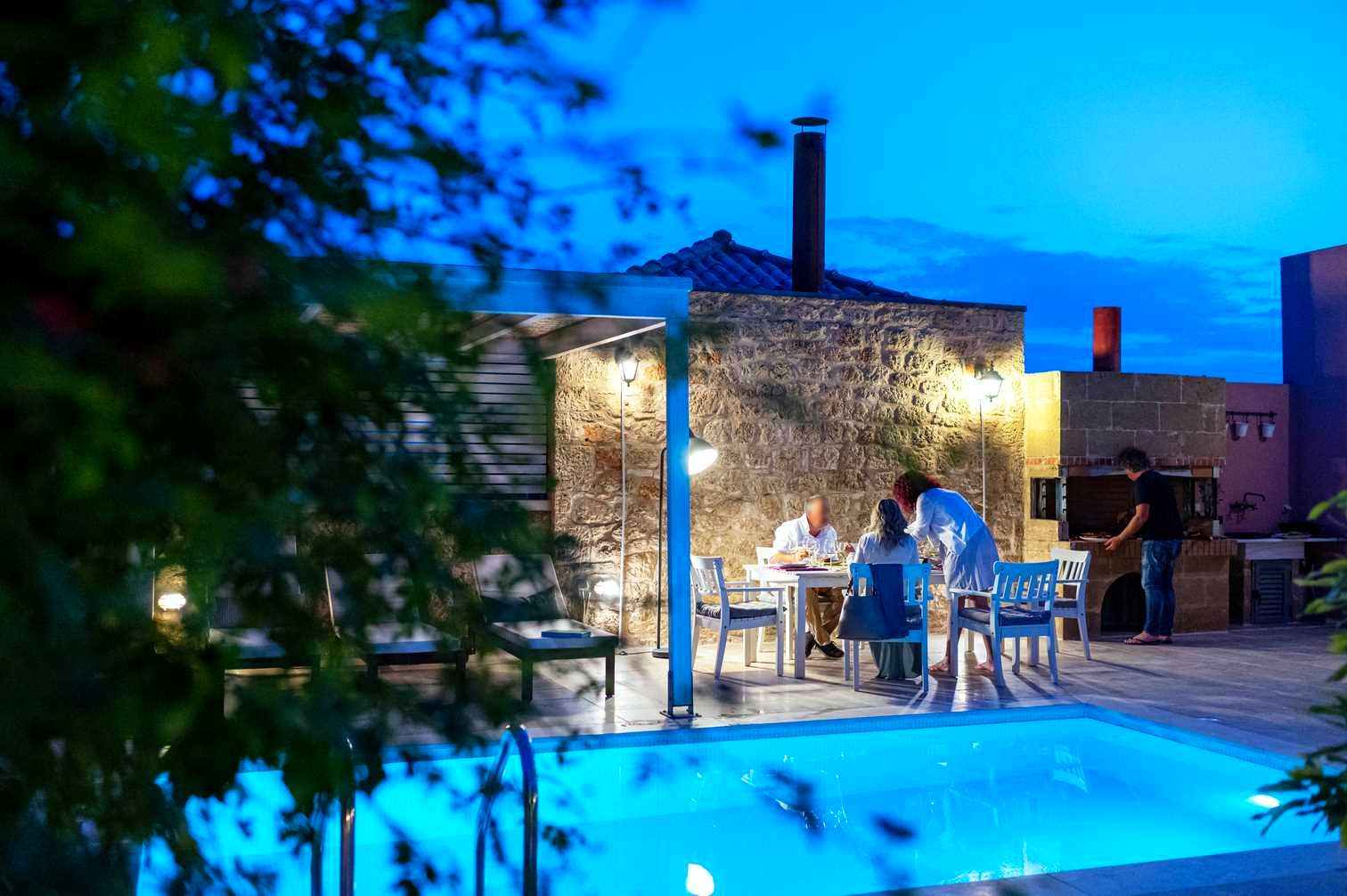 Two people sit at a white table on a patio next to a stone building. The foreground features dark foliage and a bright blue swimming pool.