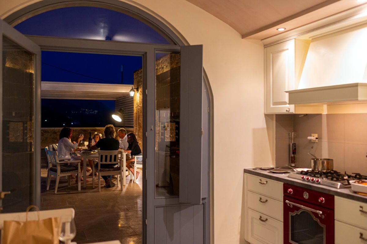 Open doorway leads to a covered patio area with a dark blue ceiling. Several people are seated at a table outdoors. Inside, a kitchen counter features a red stove and stainless steel fixtures.