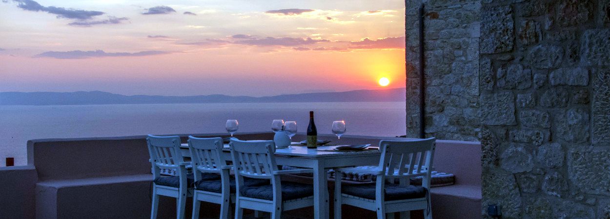 White outdoor table and four chairs are set on a patio with a view of the Aegean Sea at sunset. The sky displays shades of orange and pink near the horizon.