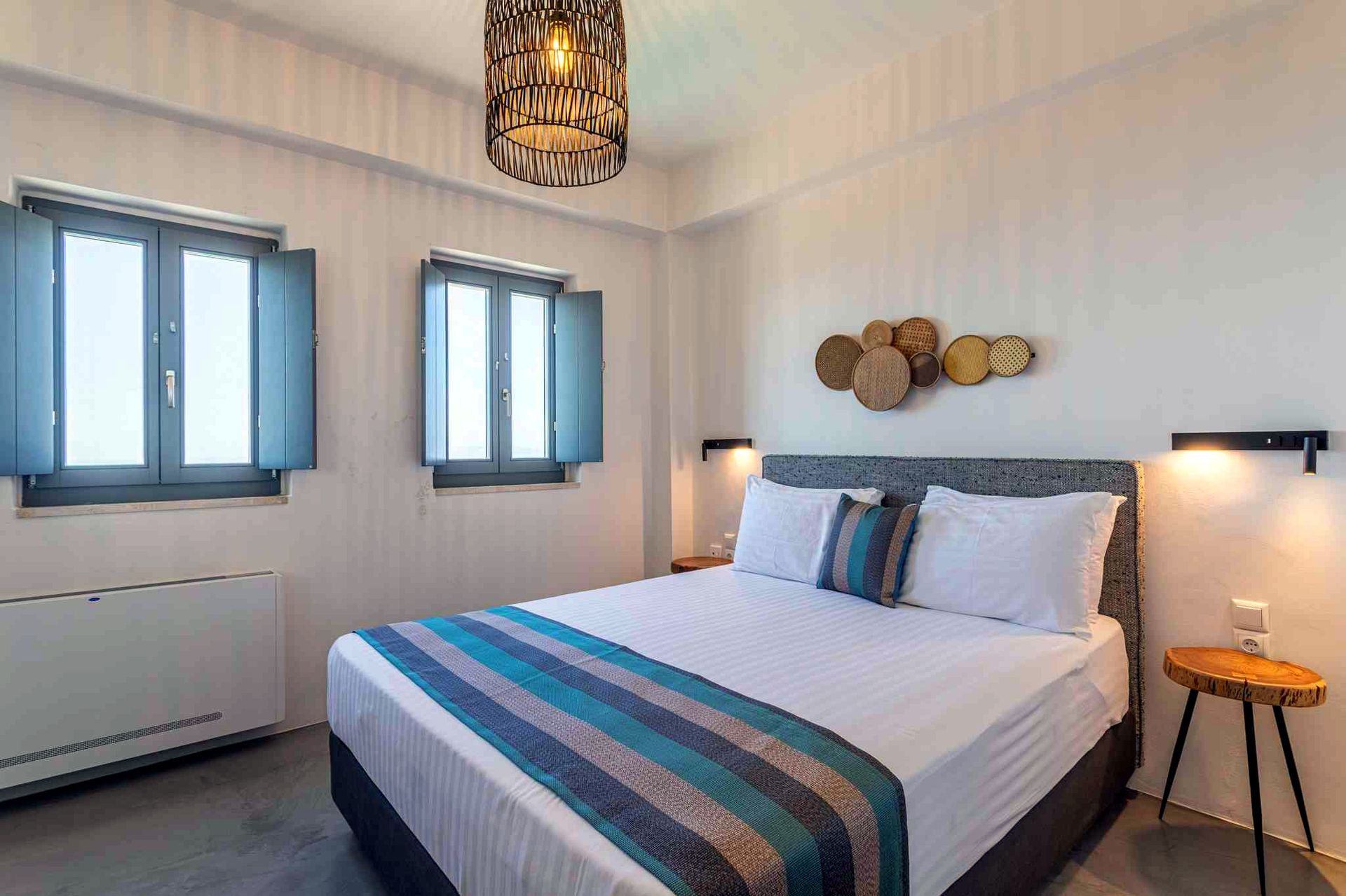 A bed with a gray headboard and white linens is centered in a room. A striped blue and gray throw is laid across the foot of the bed. To the left, two windows with light blue frames are visible, and a radiator is mounted on the wall. Above the bed, a grouping of round, light-colored decorative plate