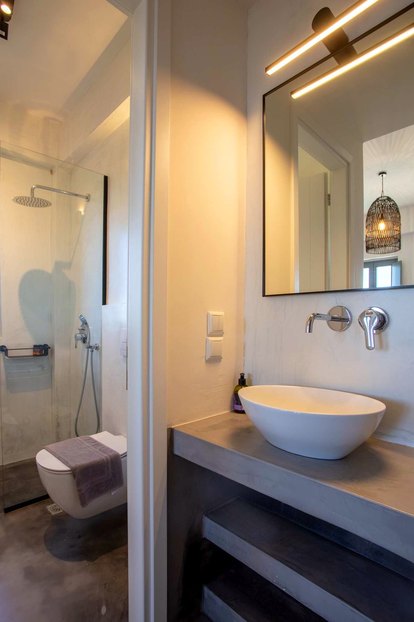 White countertop basin sits on a gray surface beneath a large, rectangular mirror illuminated by horizontal strip lights. Chrome fixtures are mounted on the wall beside the basin.