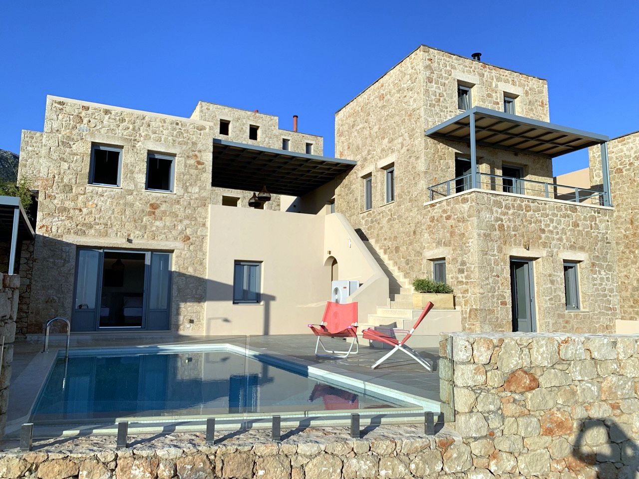 Two-story stone villas with tiled roofs and balconies overlook a rectangular swimming pool. The foreground features a low stone retaining wall.