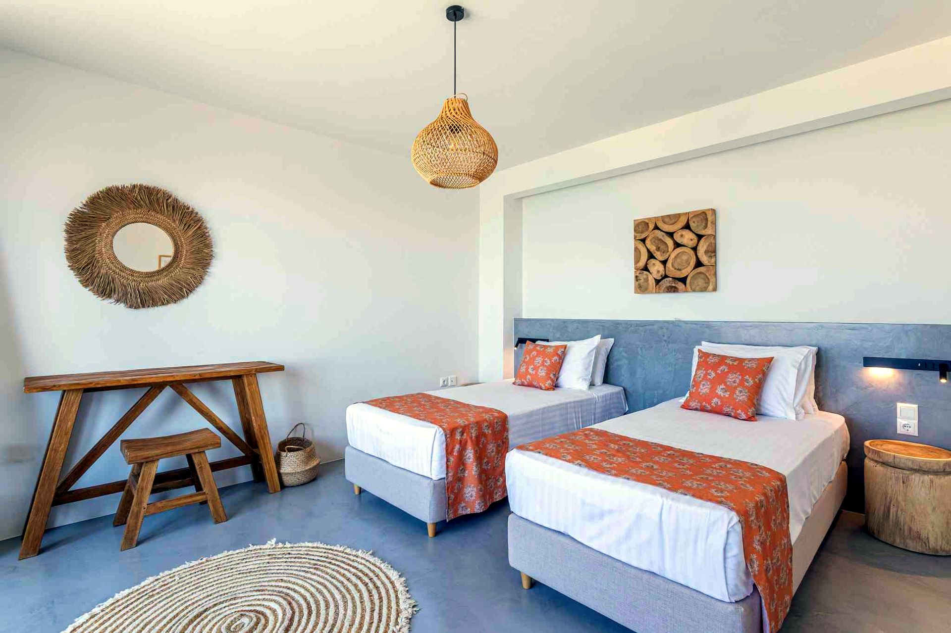 Two queen beds with white linens and orange patterned runners are positioned against a light gray wall. Above the beds, a rectangular arrangement of stacked wood slices serves as wall art. To the left, a round woven mirror hangs above a dark wood console table with a matching stool.