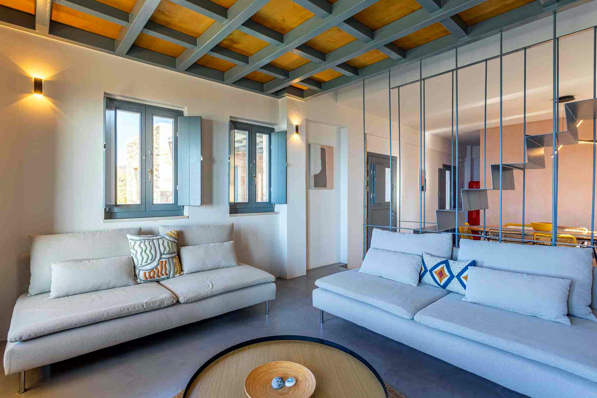 {
  "headline": "Sloped wooden ceiling structure",
  "subheadline": "Built-in seating lines the wall beneath the exposed beams and window units.",
  "alt_text": "Two light gray sofas are arranged in a room with a sloped ceiling featuring exposed wooden beams. Windows with light blue trim are visible