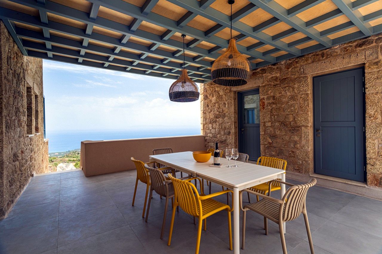 Outdoor patio with a white rectangular table and several chairs set against a textured stone wall. The patio is covered by a wooden pergola structure, and the background shows a view of the blue sea and distant land.