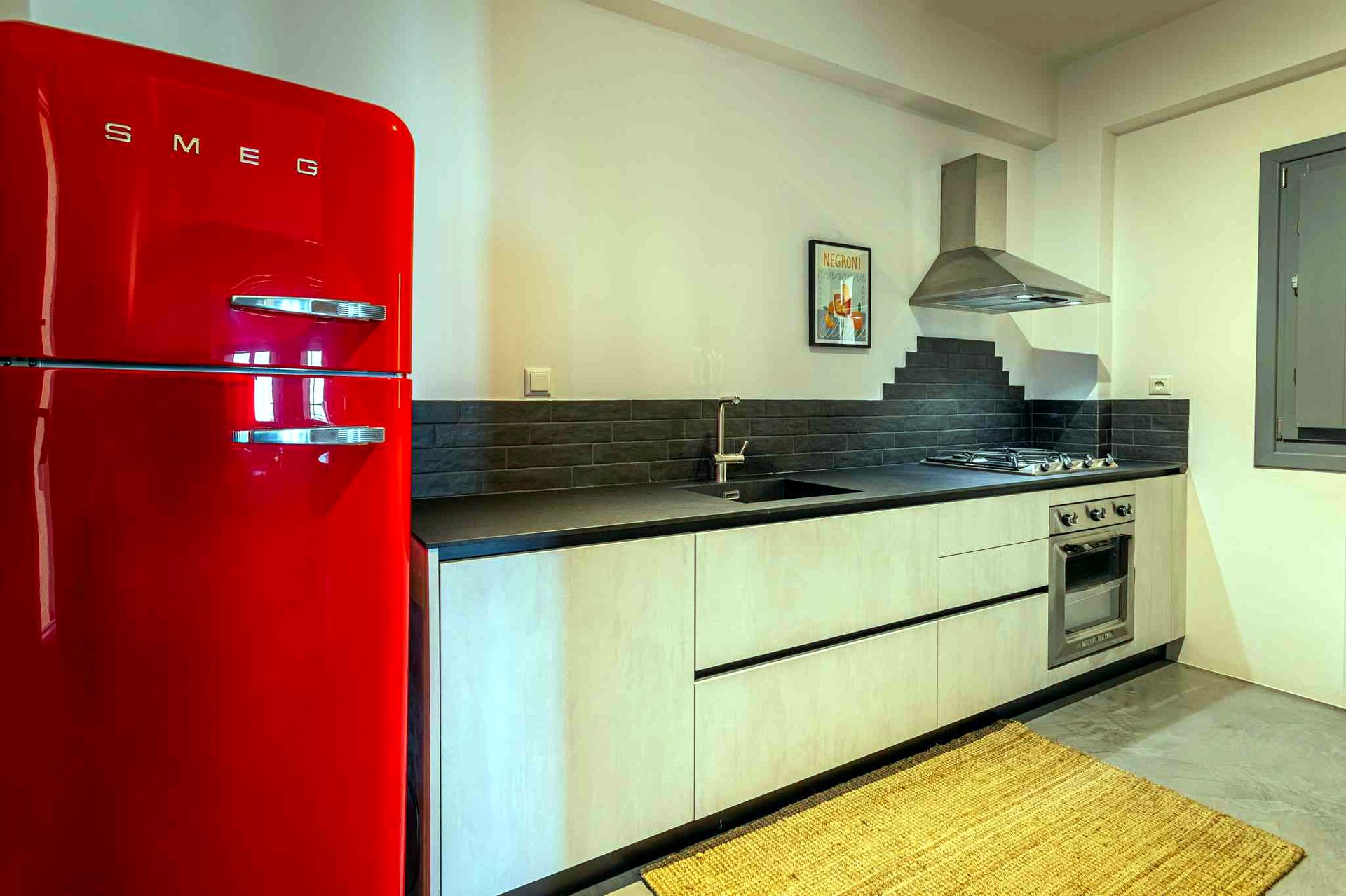 A bright red SMEG refrigerator stands next to a kitchen counter with dark gray backsplash tiles. The counter features a built-in cooktop and oven unit under a silver range hood.