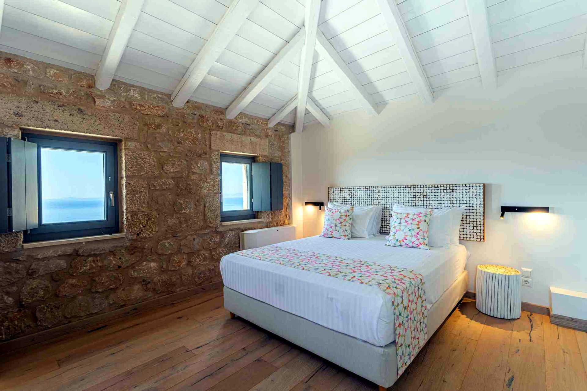 A bedroom with exposed stone walls and a pitched white beamed ceiling. Two windows are visible, one showing a view of the sea. A large bed with white linens and a patterned throw rests against the far wall.