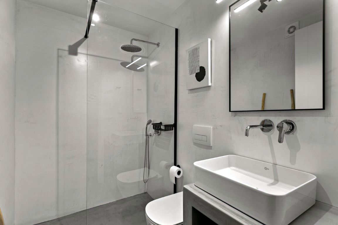 Glass shower enclosure separates a wet area from a bathroom space. Chrome shower fixtures, including a rainfall head and handheld sprayer, are mounted on the wall. A white vanity basin sits on a dark counter beneath a large rectangular mirror.
