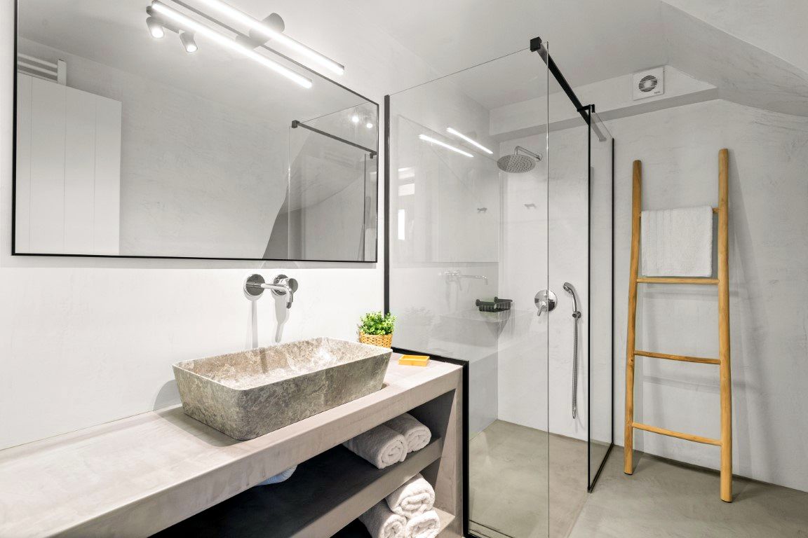 Large rectangular mirror hangs above a light-colored counter holding a textured, rectangular vessel sink. To the right, a glass shower enclosure contains a wooden ladder towel rack.