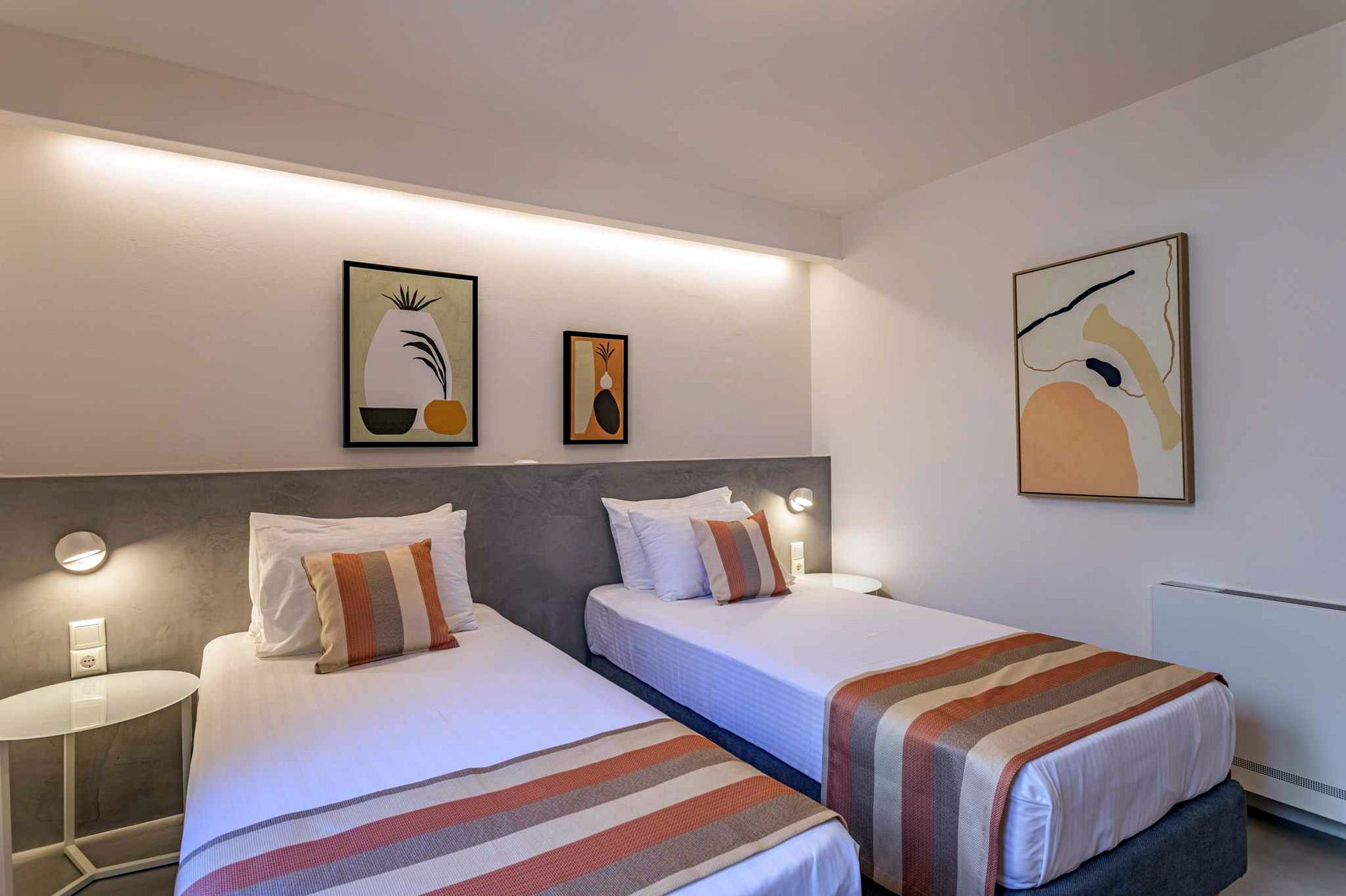 Two twin beds are set against a gray upholstered wall. Each bed has white linens and a striped throw blanket. Artwork featuring black and yellow accents hangs above the beds.
