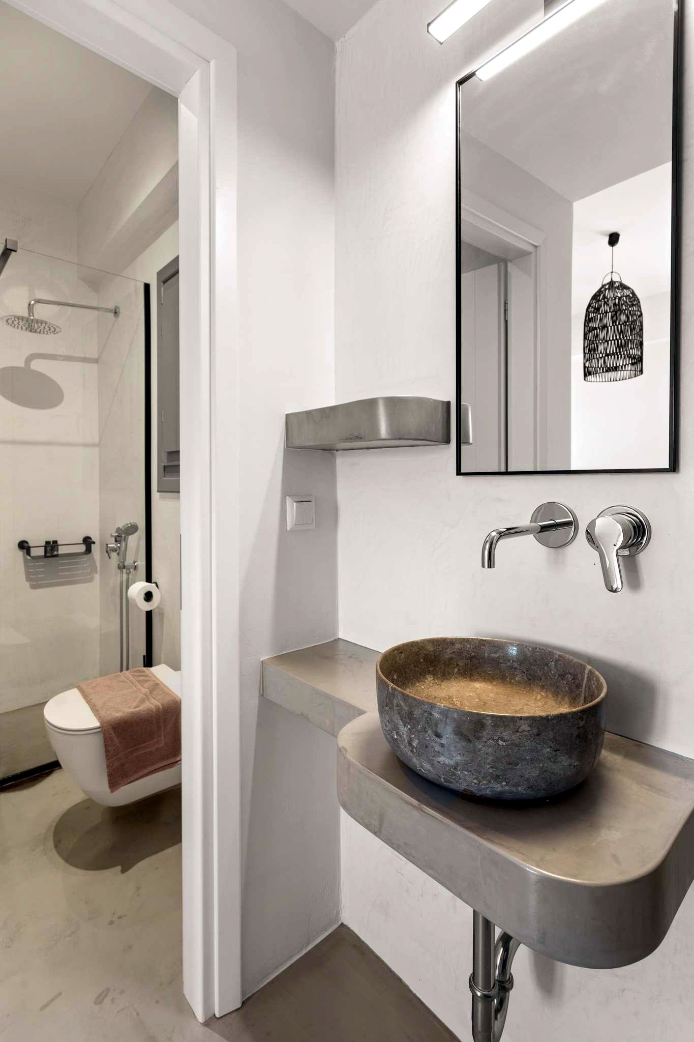 A bathroom vanity features a rectangular concrete countertop supporting a round, dark gray vessel sink. Above the sink, a large, vertical mirror hangs, reflecting a hanging black object.