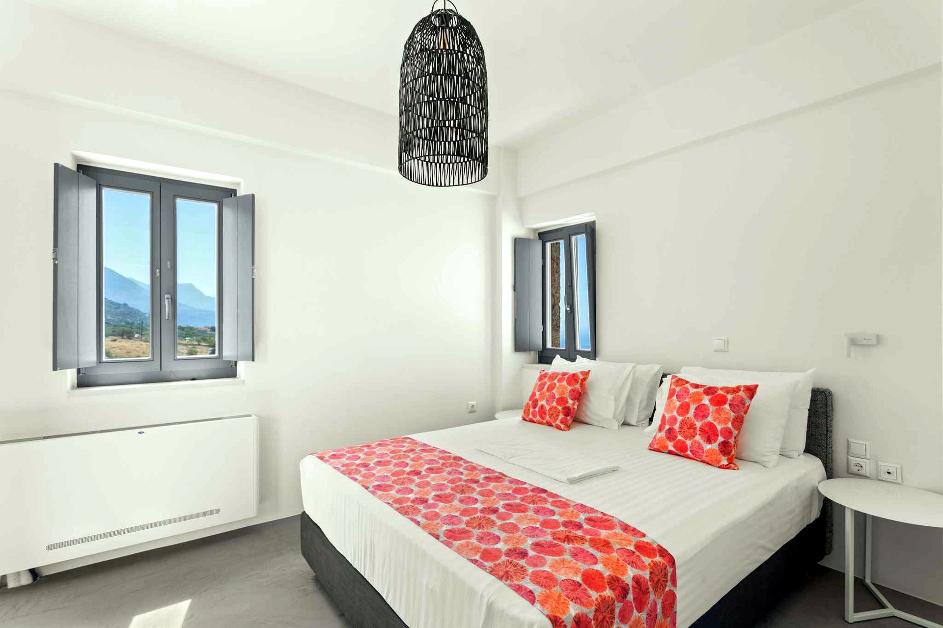 A bedroom featuring a window with two vertical panes, showing a view of blue water and distant hills. A bed with white linens and a patterned throw is centered against a white wall. A dark, woven pendant light fixture hangs from the ceiling.
