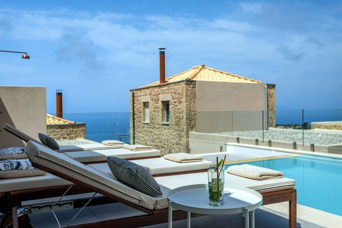 Two lounge chairs with striped cushions and one solid cushion are positioned on a patio overlooking a bright blue sea. A small white circular table holds a glass of water and a small plant.