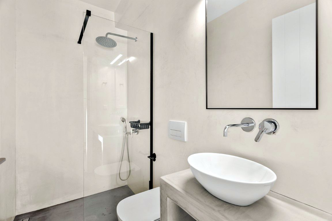 Glass shower enclosure features a rainfall shower head and handheld fixture. A white sink basin rests on a light-colored countertop below a rectangular mirror.