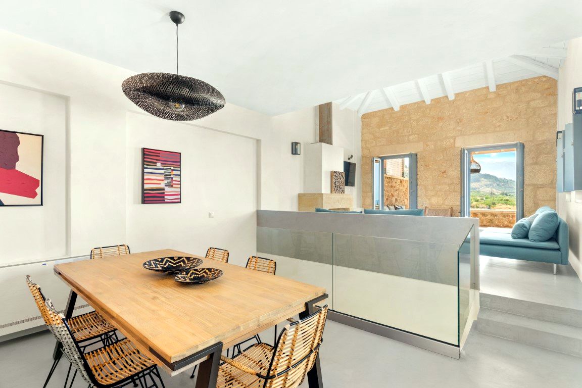 Large wooden dining table with woven chairs sits in a bright room with white walls and a vaulted ceiling. A low counter separates the foreground from a seating area with blue cushions and a view through glass doors.