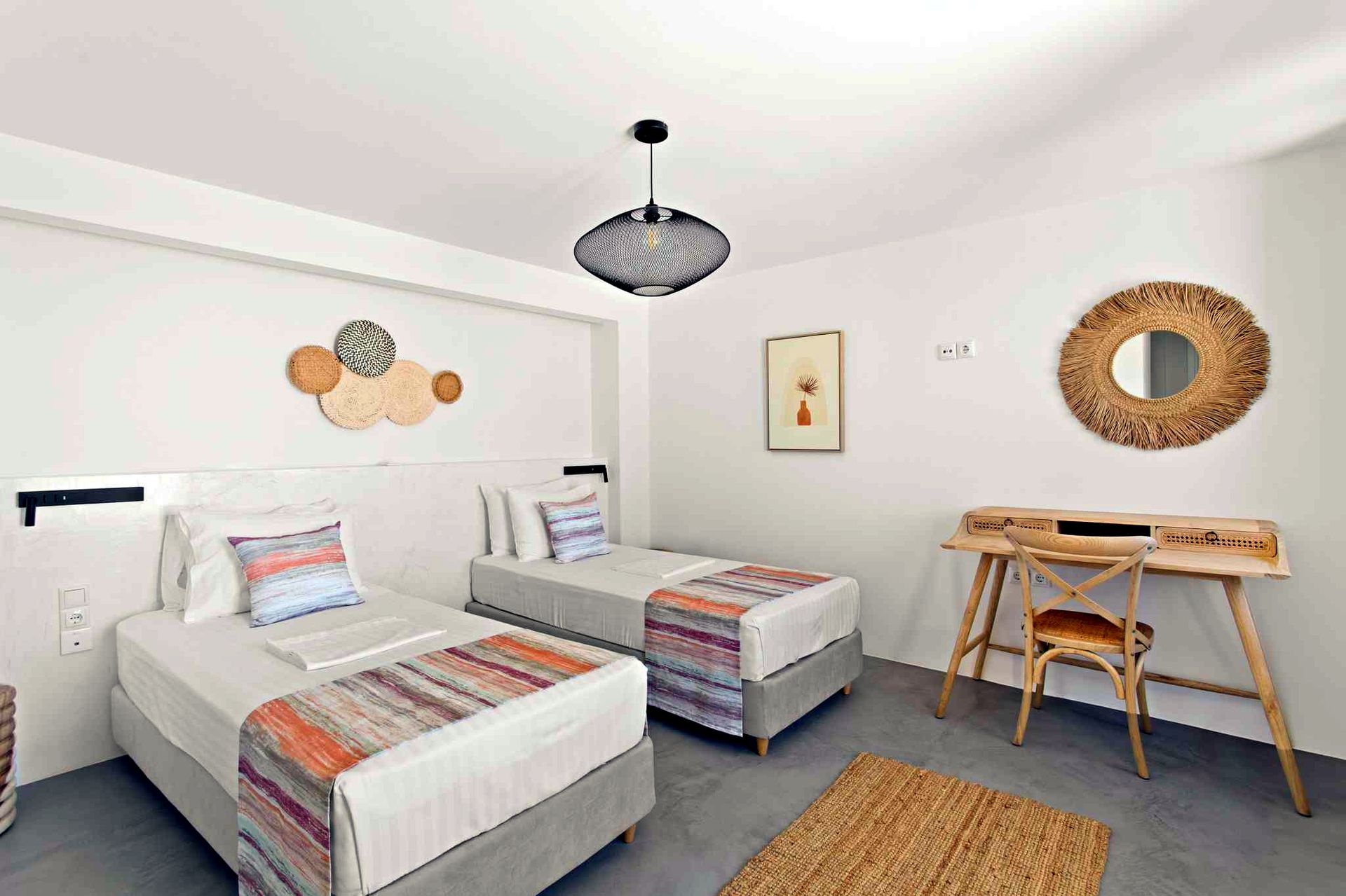 Two twin beds with striped throw blankets are positioned against a white wall. Above the beds, three circular decorative elements are mounted. To the right, a wooden console table holds a mirror and a small framed picture.