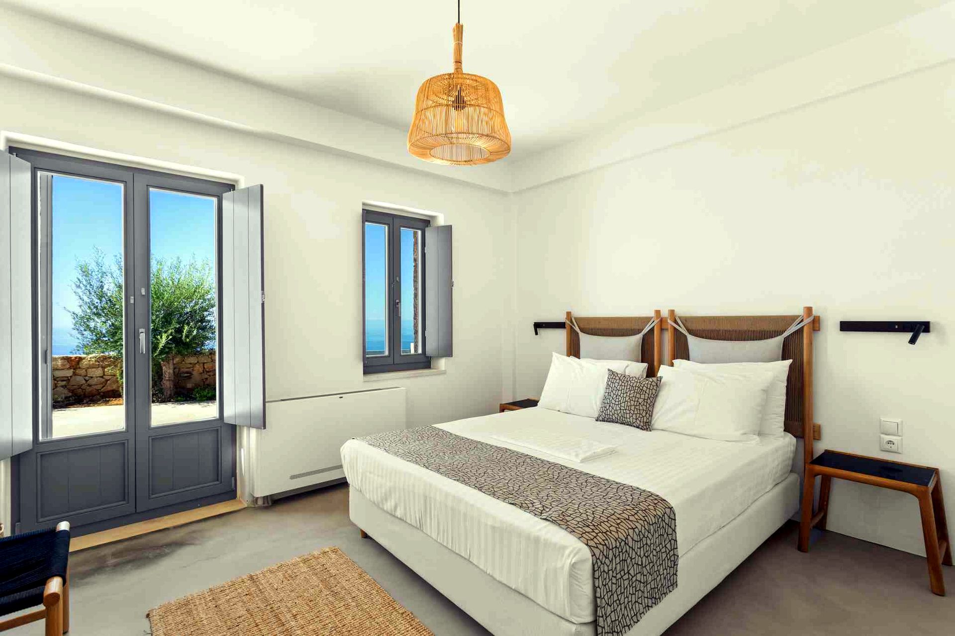Two sets of sliding glass doors open onto a patio overlooking the sea. The room contains two twin beds with white linens and a woven pendant light fixture.