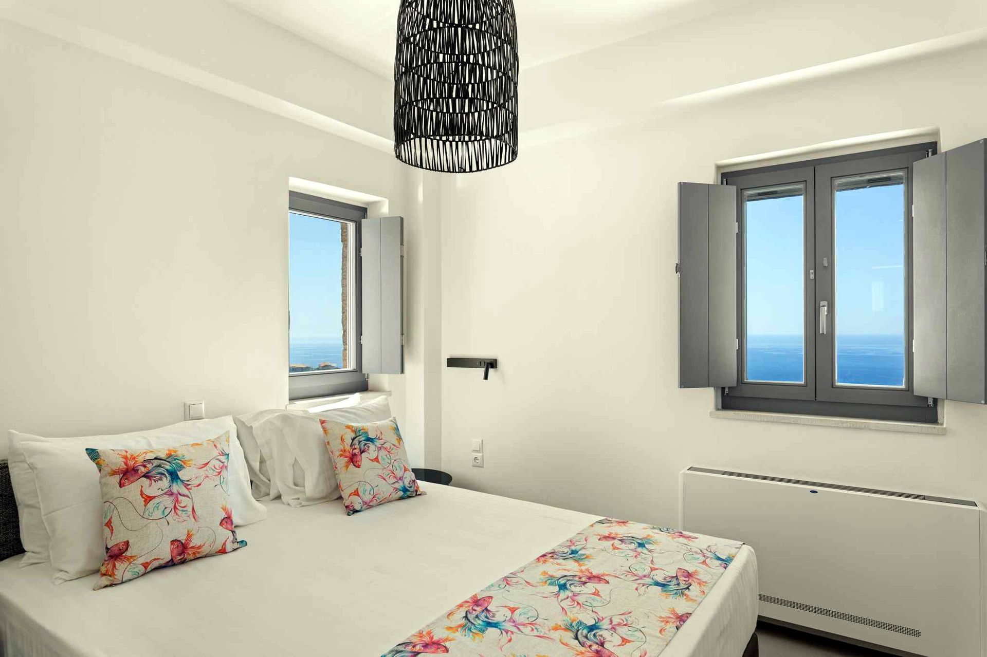 A bedroom with white bedding and patterned pillows faces a window showing a view of the blue Aegean Sea. The room has light gray trim and a dark woven pendant light hangs from the ceiling.