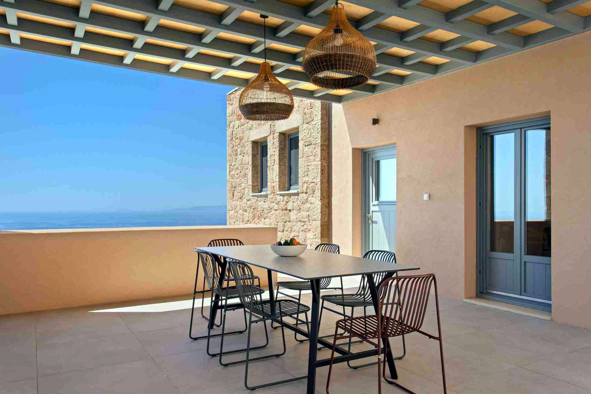 Outdoor patio with a rectangular table and chairs set against a backdrop of blue sea and sky. Three woven pendant lights hang from a slatted overhead structure.