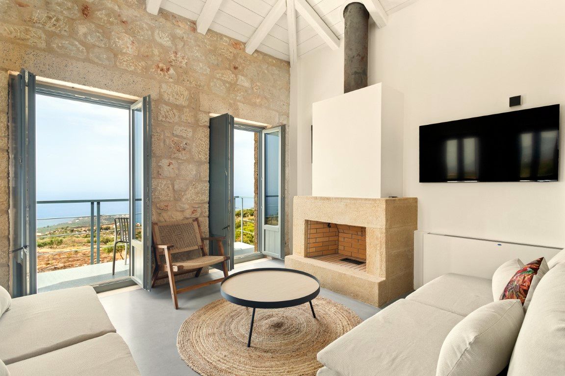 Large sliding glass doors open onto a balcony with metal railings overlooking the sea. The interior features exposed stone walls, a white fireplace structure, and a low circular coffee table on a woven rug.