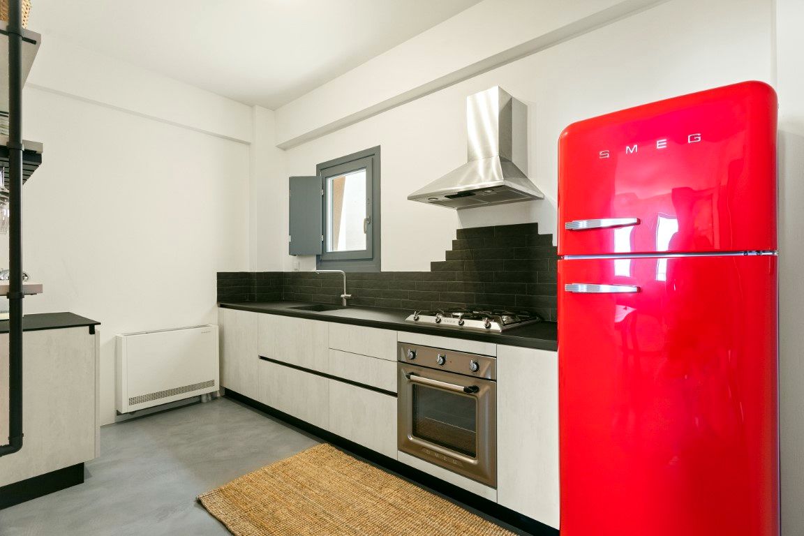 Stainless steel range hood hangs over a black cooktop set into white cabinets. A red refrigerator stands to the right, next to a sink area with a window.