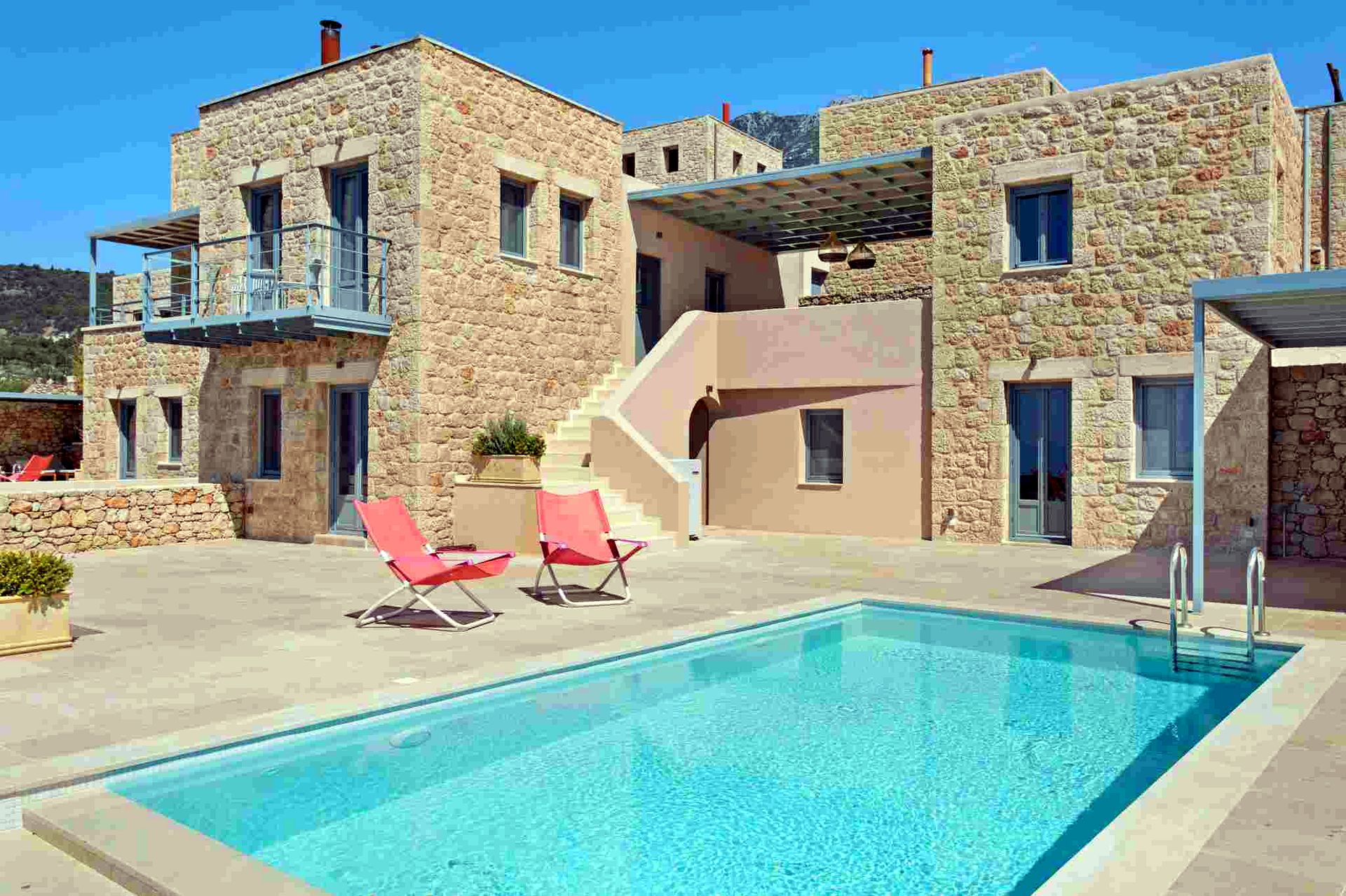 Two-story stone villa complex featuring multiple windows and balconies overlooks a rectangular blue swimming pool. Red and white lounge chairs are placed on the surrounding paved patio.
