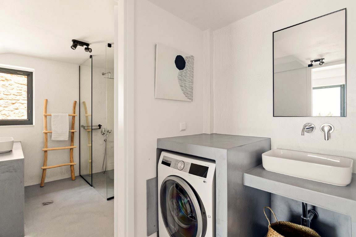 White bathroom interior featuring a stacked washer and dryer unit beneath a gray countertop. A rectangular white sink basin sits on the counter, and a large mirror hangs above it. A towel rack stands against the far wall.