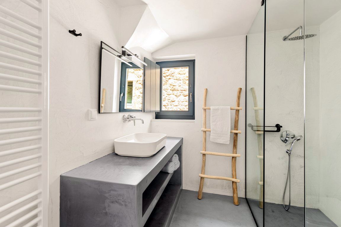 White rectangular basin sits on a gray concrete vanity counter beneath a mirror. A window with light-colored stone visible outside is centered above the sink area.