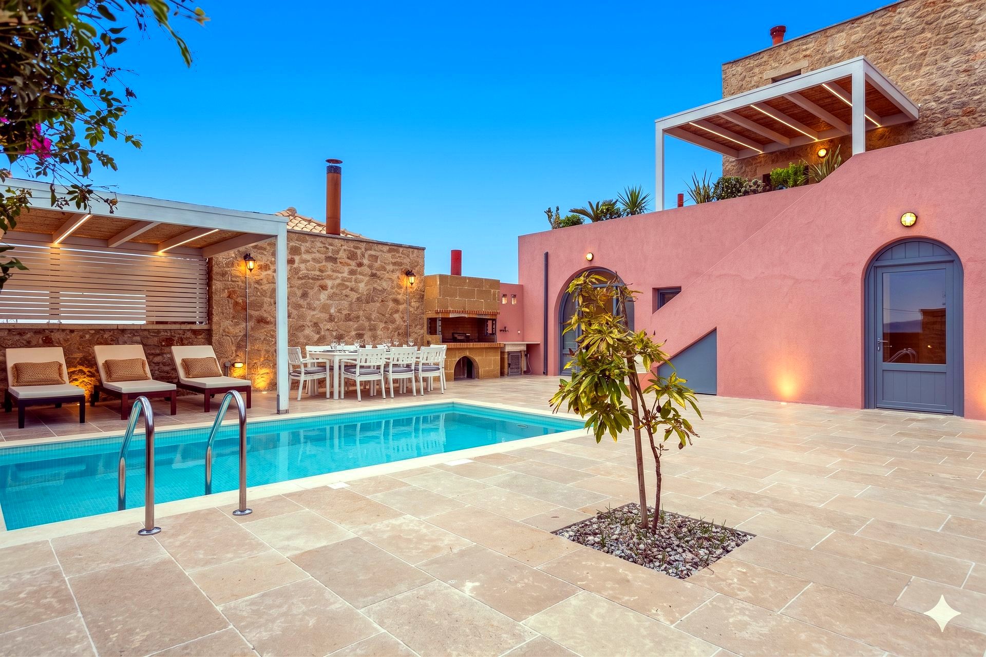 A large tiled patio surrounds a rectangular blue swimming pool. To the right is a pink stucco building with a covered balcony and a blue door. Outdoor furniture and a dining table are visible.